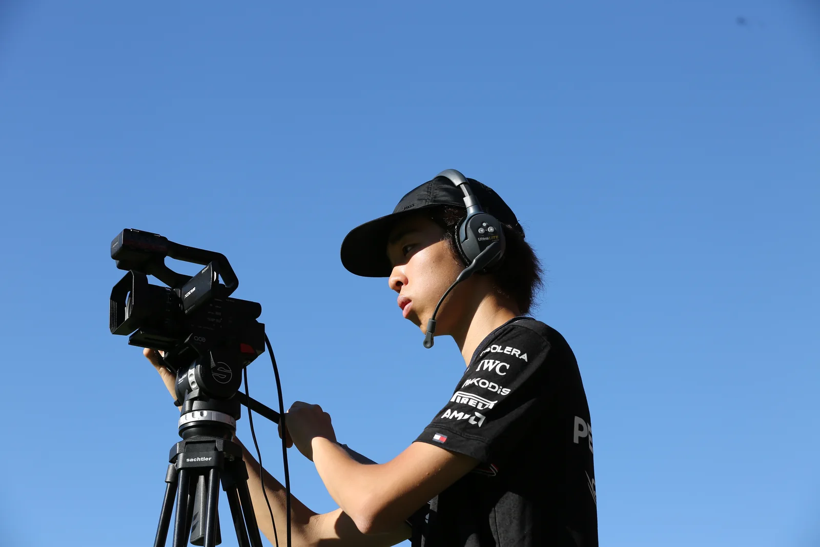 A student camera operator filming outdoors with a tripod-mounted video camera and headset